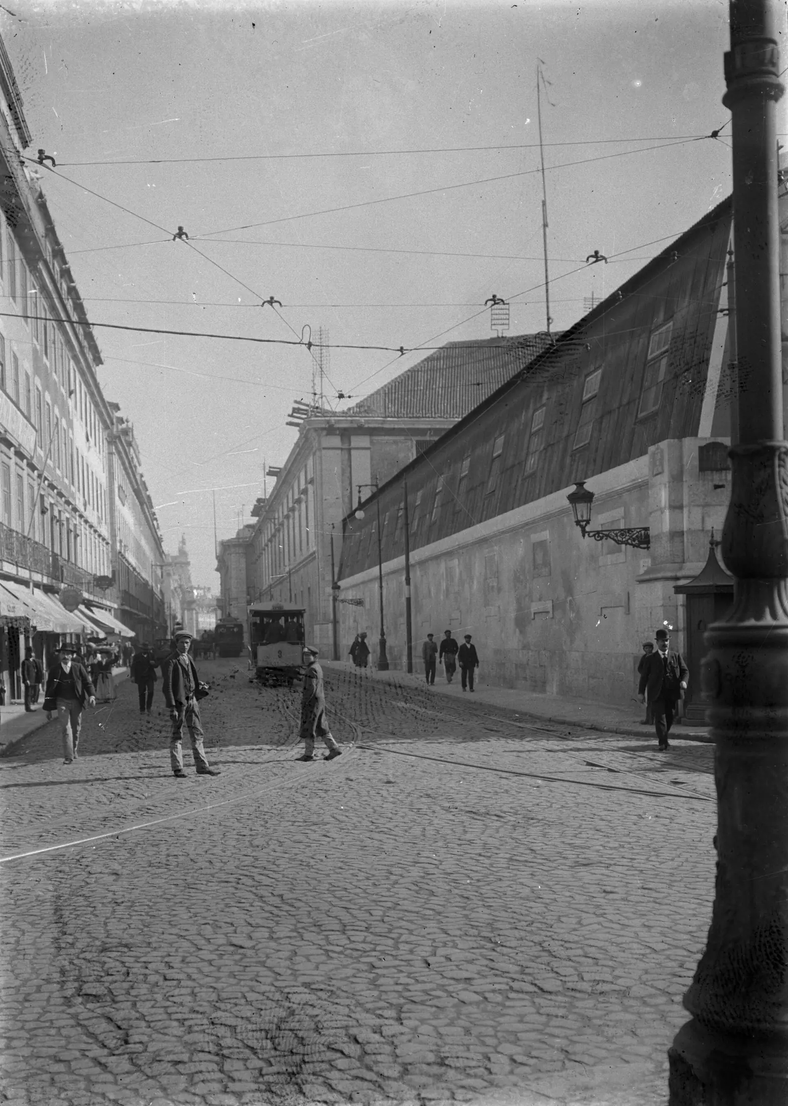 Fotografia a preto e branco de uma rua empedrada com carris e candeeiros, um muro comprido à direita e algumas pessoas ao fundo.