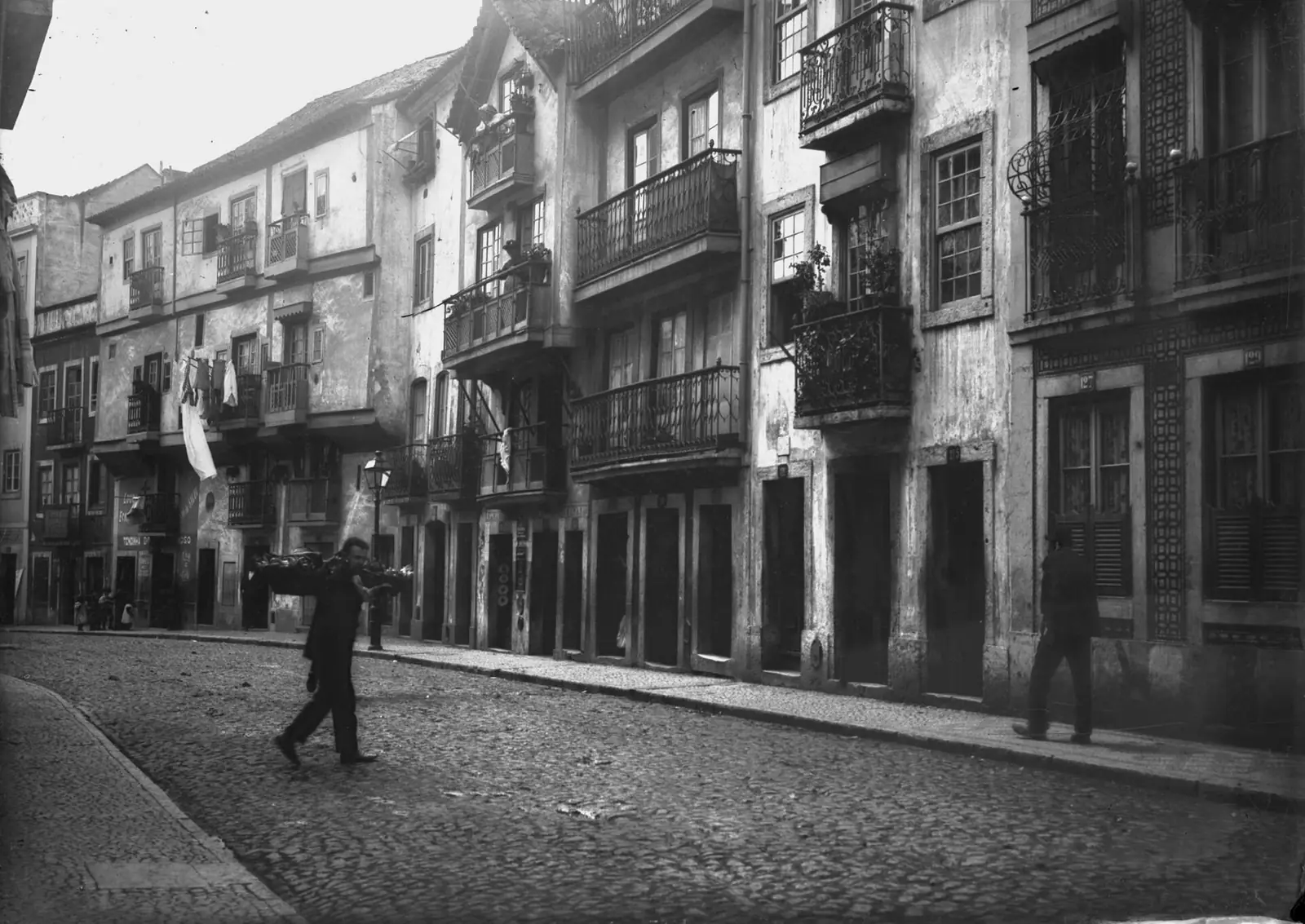 Fotografia a preto e branco de uma rua empedrada entre prédios antigos de varandas de ferro, com um homem a atravessar.