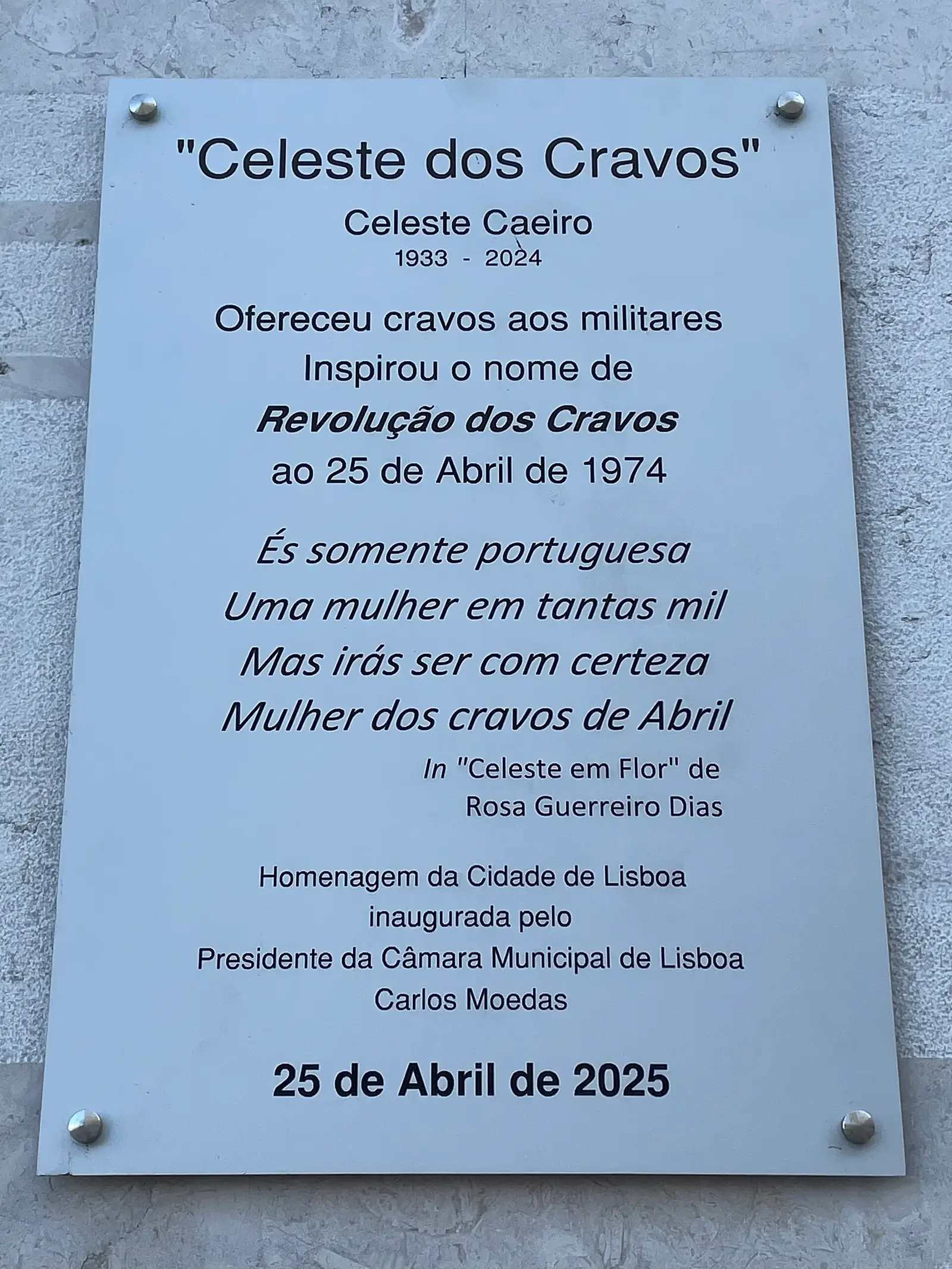 Placa metálica clara com texto gravado: o nome de Celeste Caeiro, as datas 1933–2024, uma frase sobre os cravos oferecidos aos militares e uma quadra em itálico.