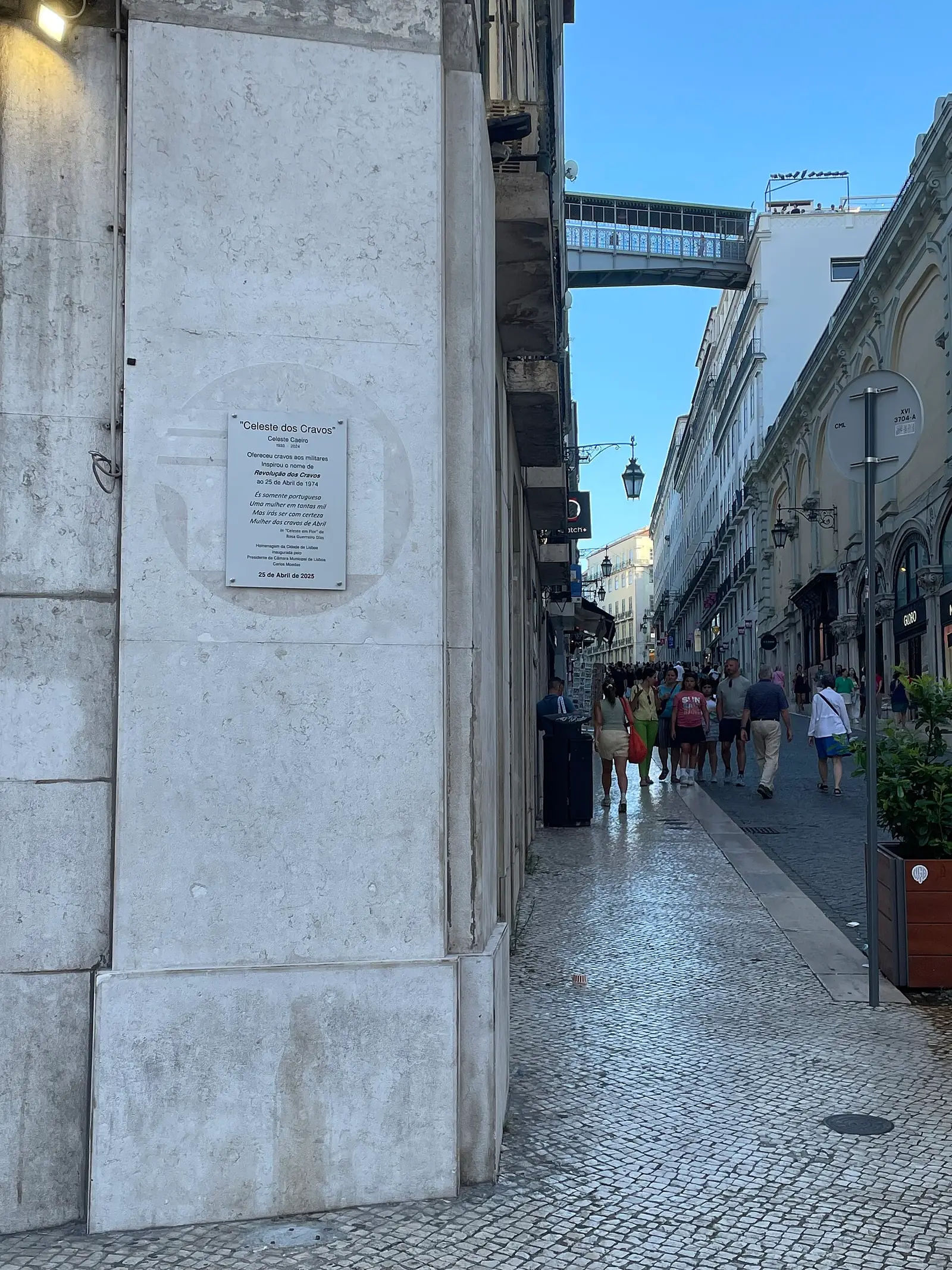 Esquina de pedra lisa com uma pequena placa clara à altura dos olhos; à direita, a rua estreita sobe entre fachadas, com uma ponte metálica ao fundo.