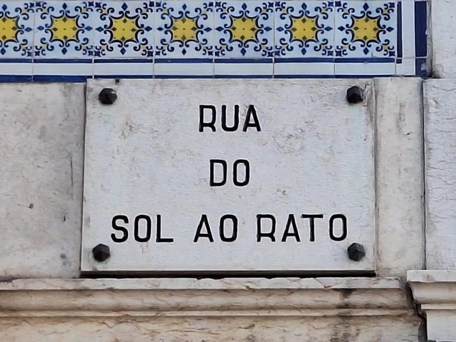 Placa de pedra clara com as palavras «Rua do Sol ao Rato» gravadas a negro, presa por quatro parafusos sob uma faixa de azulejos azuis e amarelos.