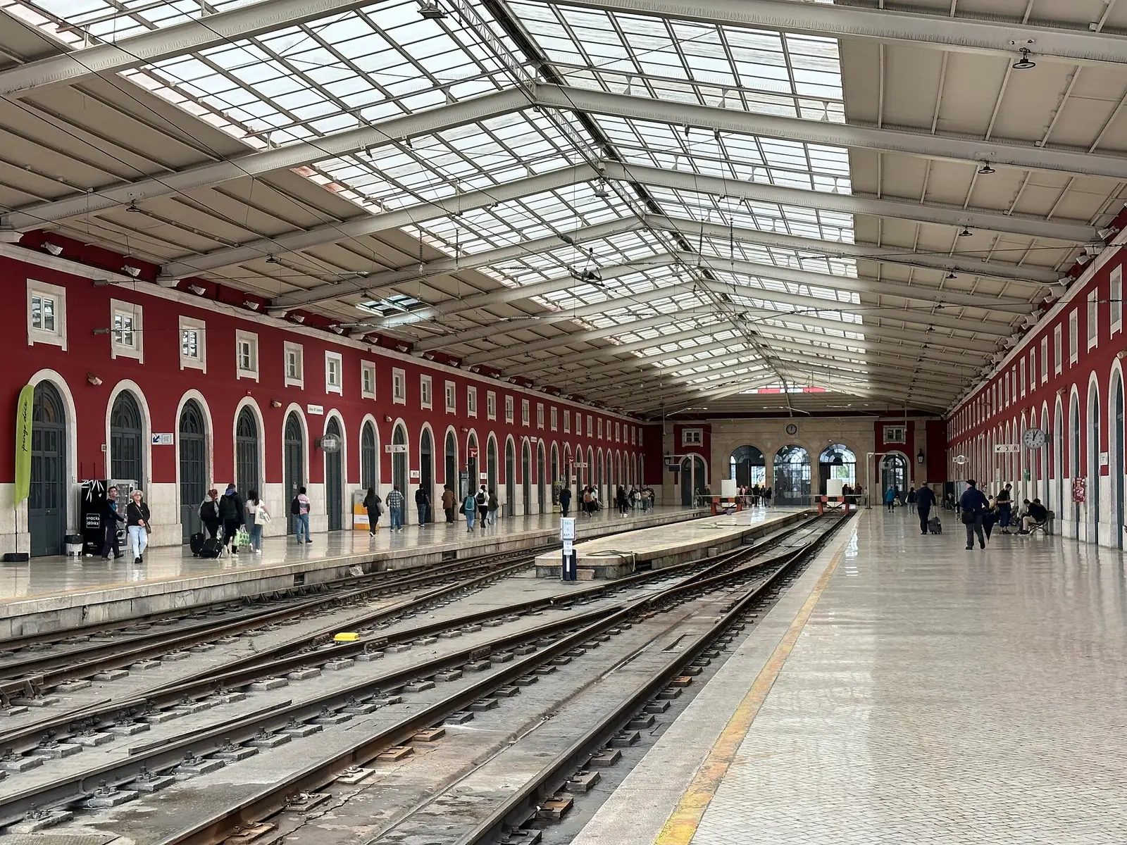 Gare comprida com telhado de vidro e ferro sobre as linhas, paredes vermelhas com arcos de um lado e passageiros no cais.