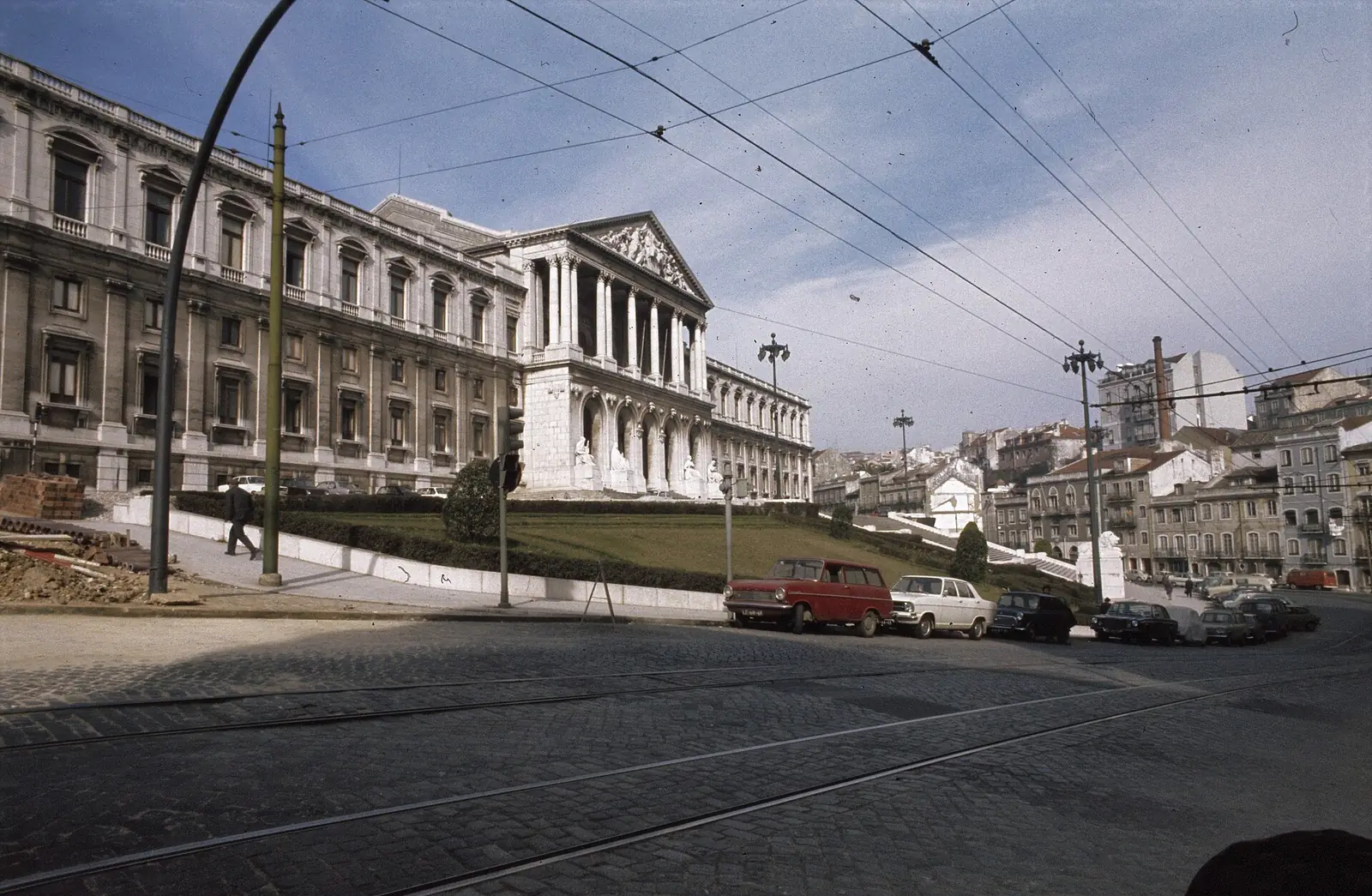 Fotografia a cores, desbotada, da fachada comprida e branca do palácio com a colunata ao centro, vista de uma rua com fios de elétrico e automóveis da época estacionados.