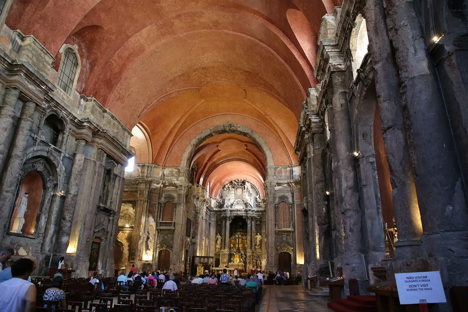Nave alta de teto abobadado cor de tijolo, com colunas e paredes de pedra escurecida e lascada, filas de bancos e o altar iluminado ao fundo.