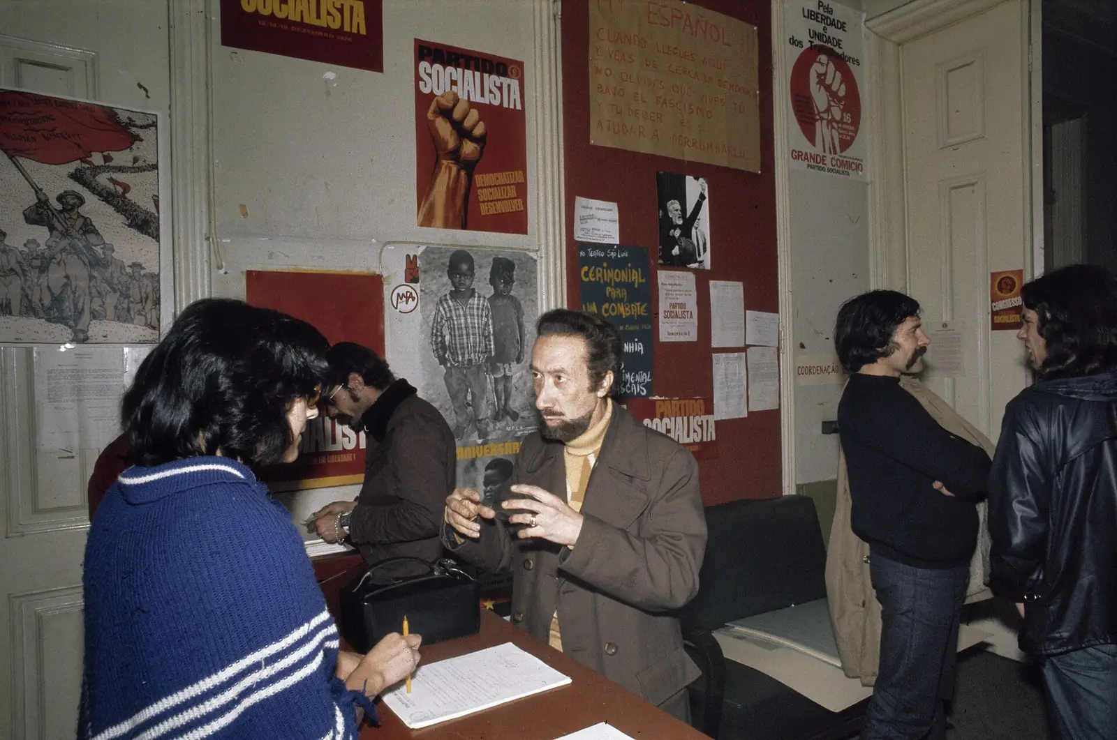 Fotografia a cores de uma sala com as paredes cobertas de cartazes, entre eles um punho erguido com as palavras «Partido Socialista»; um homem de barba fala com uma jovem sentada a uma mesa com papéis, e outros conversam ao fundo.