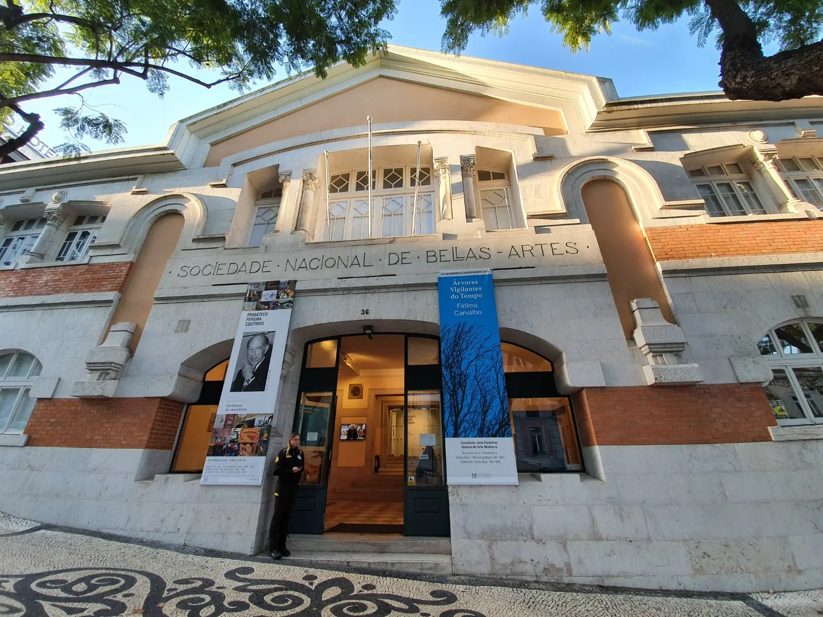 Edifício de dois pisos, de fachada clara com faixas de tijolo à vista, grandes janelas em arco e um painel azul junto à porta, sob céu azul.