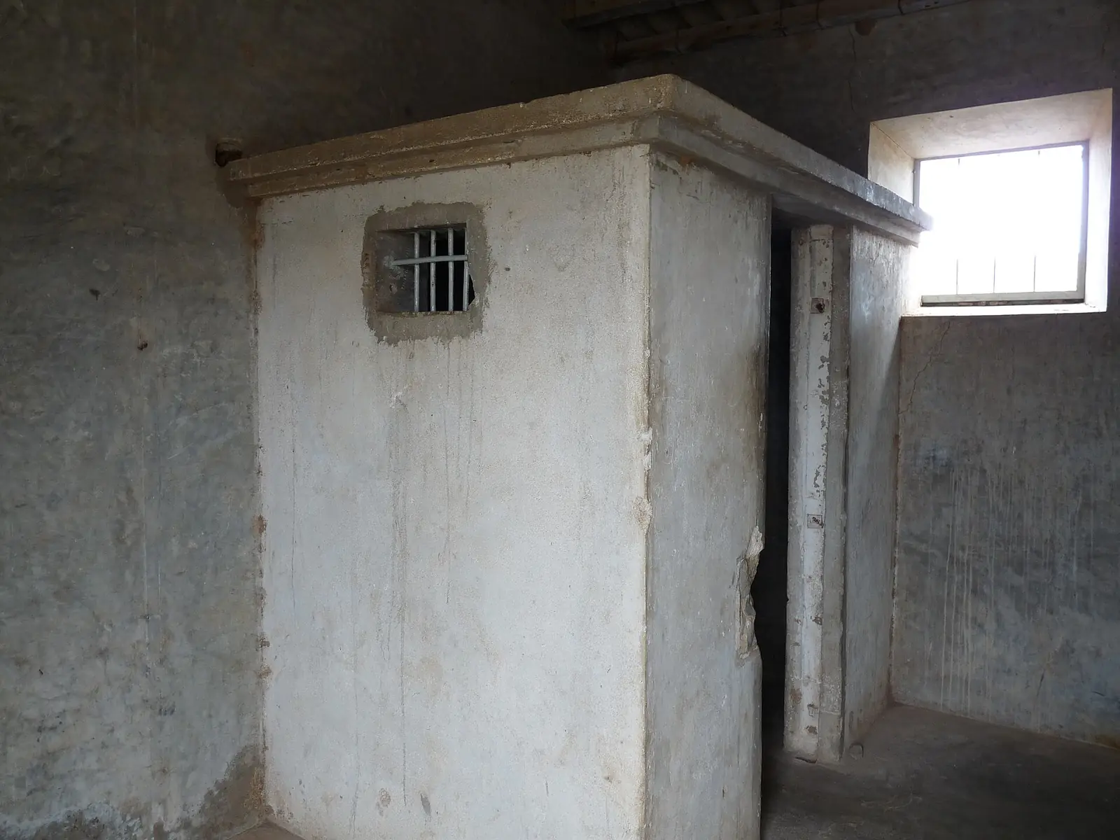 Interior de cimento nu e manchado, com um cubículo fechado de paredes grossas e uma pequena janela gradeada.