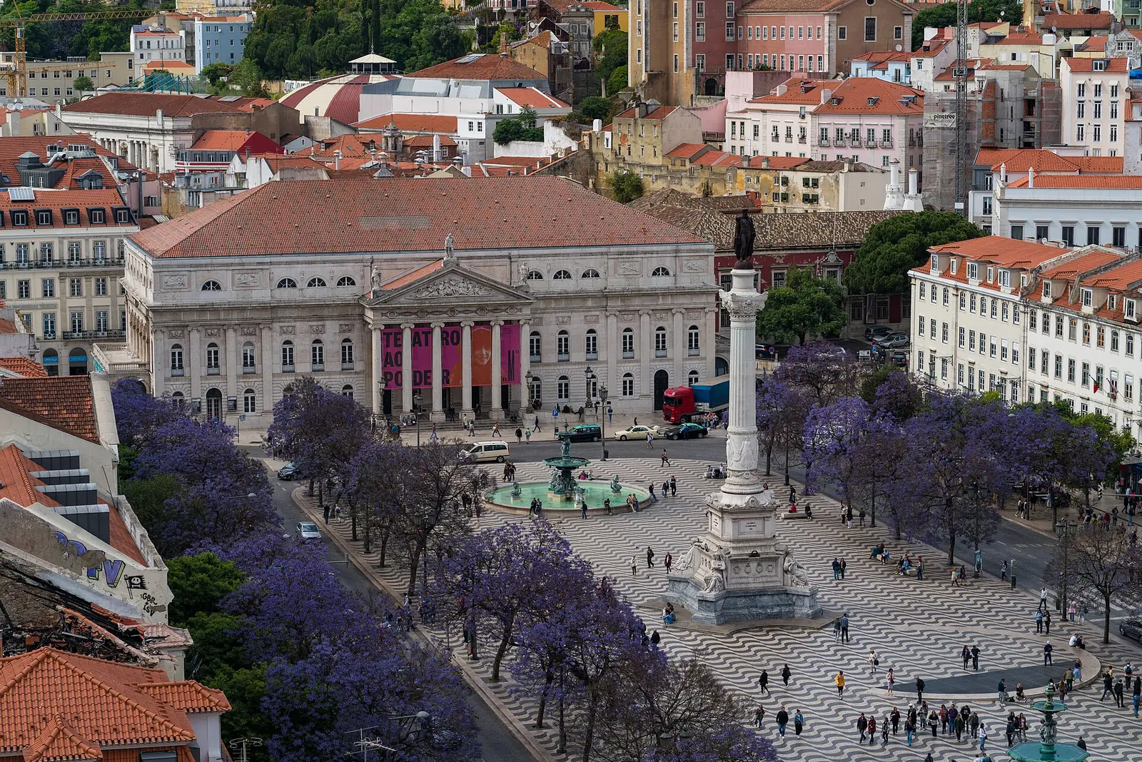 Vista de cima de uma praça grande com calçada em ondas, uma coluna alta e jacarandás floridos de lilás; ao fundo, o edifício comprido do teatro, com frontão e colunas, entre telhados vermelhos.