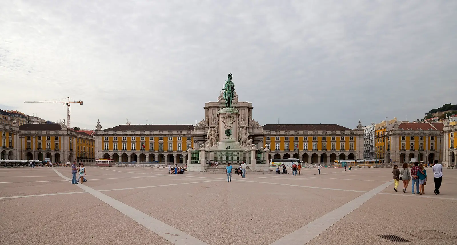 Praça larga sob céu nublado, com a estátua equestre ao centro e o arco triunfal ao fundo, entre edifícios amarelos com arcadas; algumas pessoas a passear.