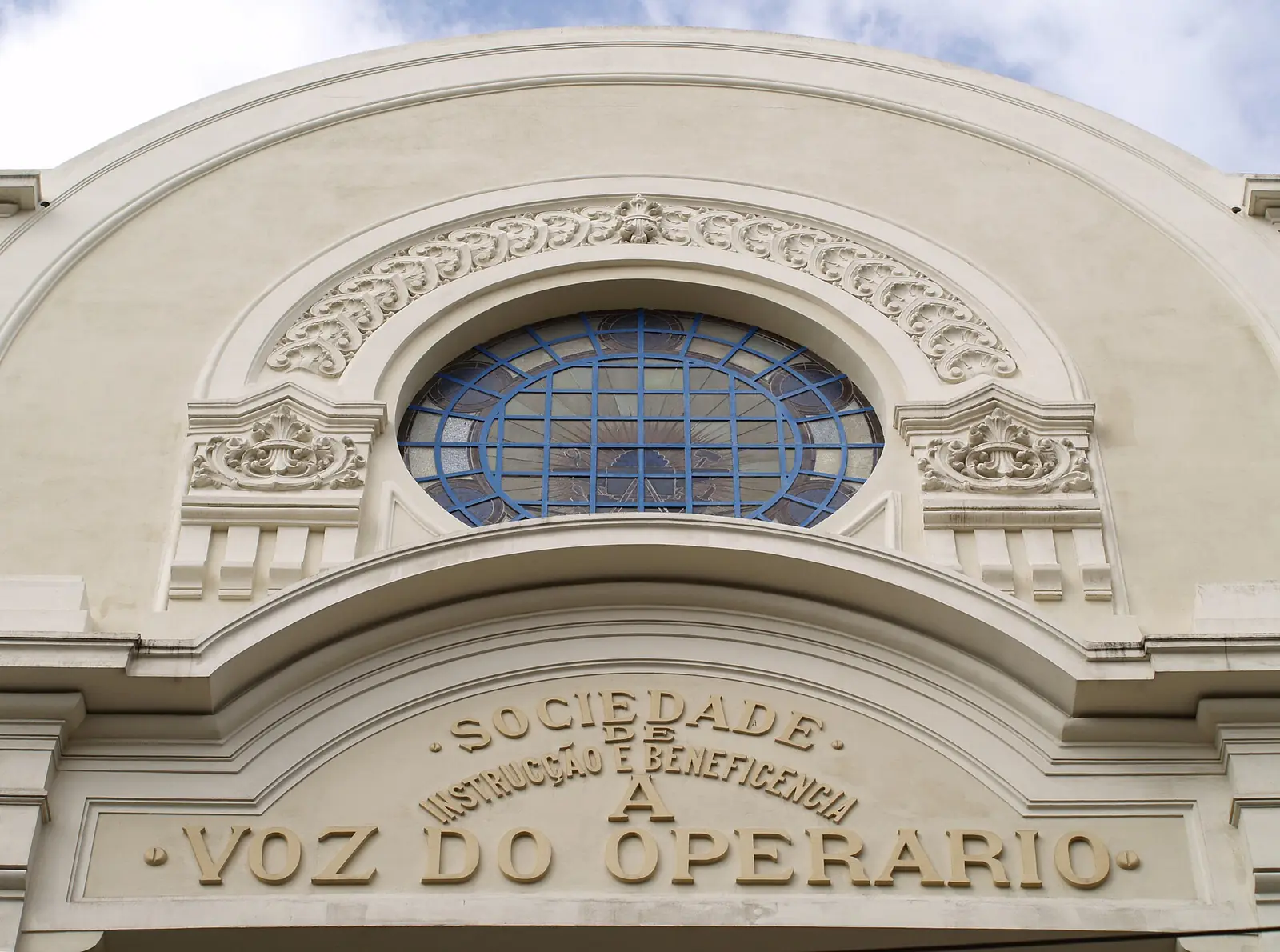 Arco de pedra clara com uma janela redonda de vidros azuis e, por baixo, o nome da sociedade em letras de relevo, a terminar em «A Voz do Operario».